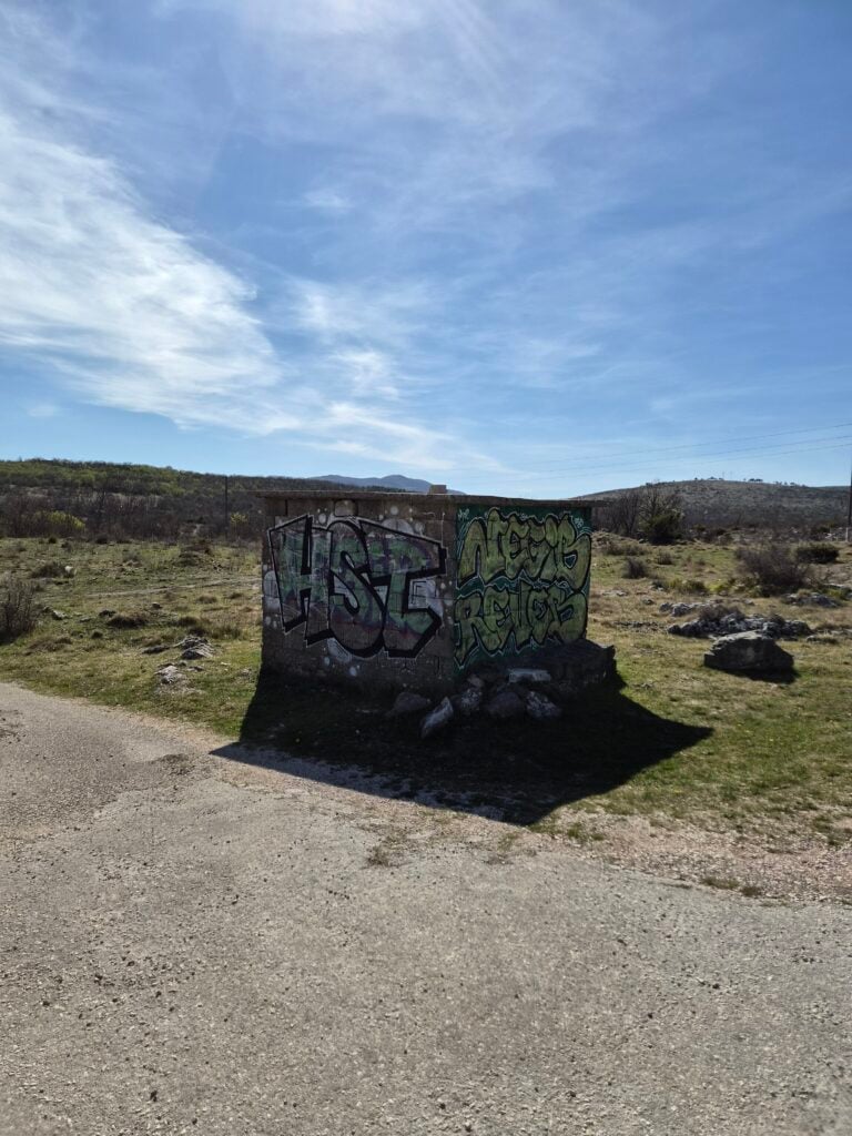 Bočni pogled na malu kamenu građevinu s manje grafita i dalekim krškim brdima, snimljeno Samsung S26 kamerom.