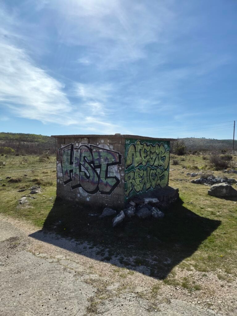 Pogled na istu malu kamenu građevinu u travnatom polju s vidljivim grafitima na jednoj strani, test Galaxy S26 pejzaža.