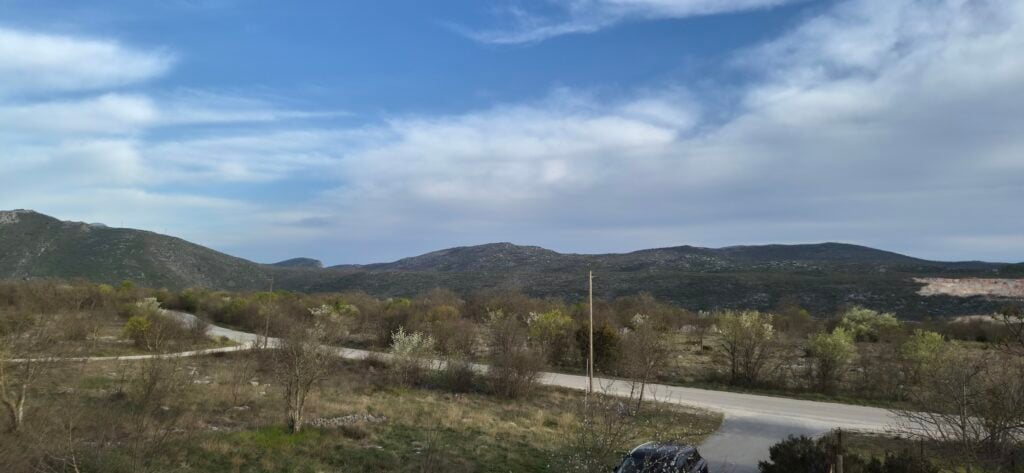 Perspektiva s razine tla na nepopločani, travnati put koji vodi u polje prema dalekim brdima, snimljeno Galaxy S26 kamerom.